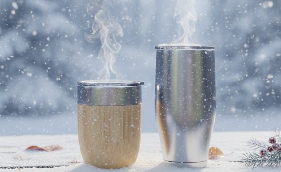 Zwei Thermobecher – einer in Holzoptik und einer aus Edelstahl – stehen auf einer verschneiten Holzbank im Freien. Aus beiden Bechern steigt deutlich sichtbarer Dampf auf, während im Hintergrund eine verschneite Winterlandschaft bei leichtem Schneefall zu sehen ist.